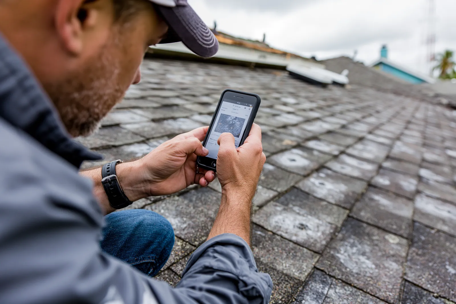 A weathered shingle roof is photographed by a roofer with a smartphone