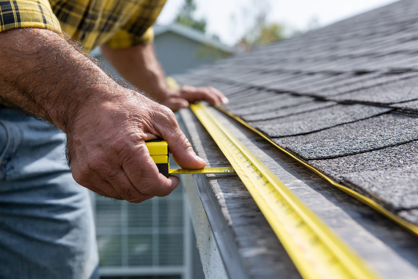 A roofer measures a roof edge using a yellow tape measure