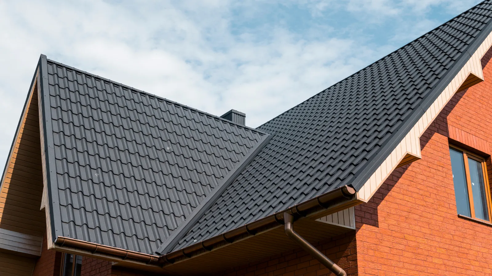 Brick house with dark metal tile roofing under a partly cloudy sky